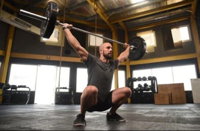 The photo focuses on a man in a cross-training studio. He is doing squats with the help of dumbbells and weights.