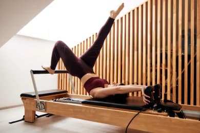 In the photo, a woman is in a pilates studio performing an exercise that also helps to stretch the legs.