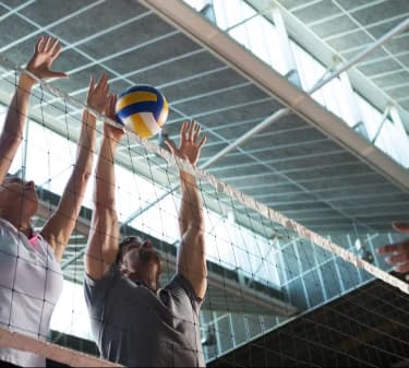 The image shows three people playing volleyball. The photo was taken in a gym, and two of the three people are blocking the ball by stretching their arms above the net.